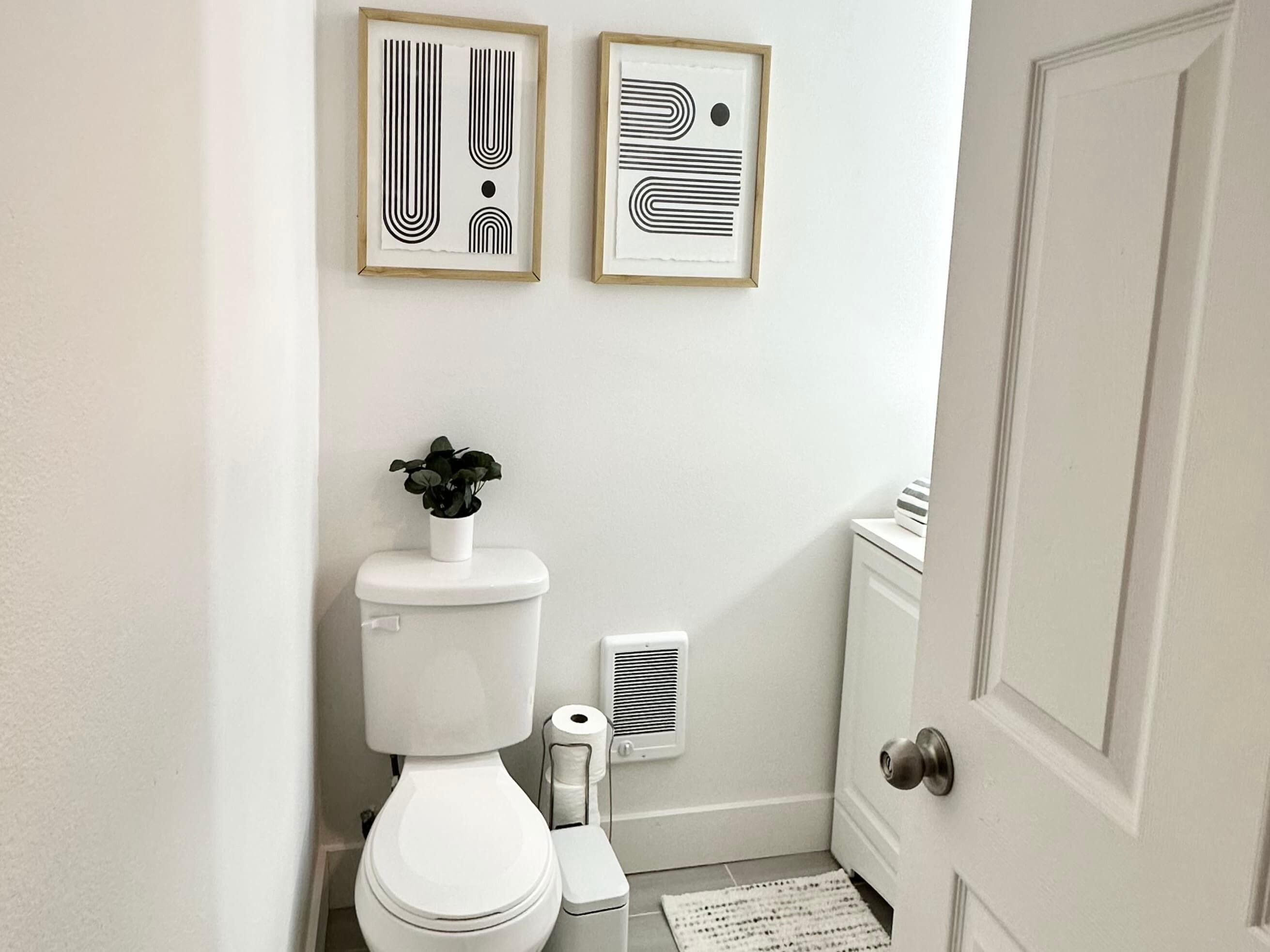 A bathroom with a toilet, a small plant on the toilet tank, a framed abstract art piece above the toilet, and a door to the right.