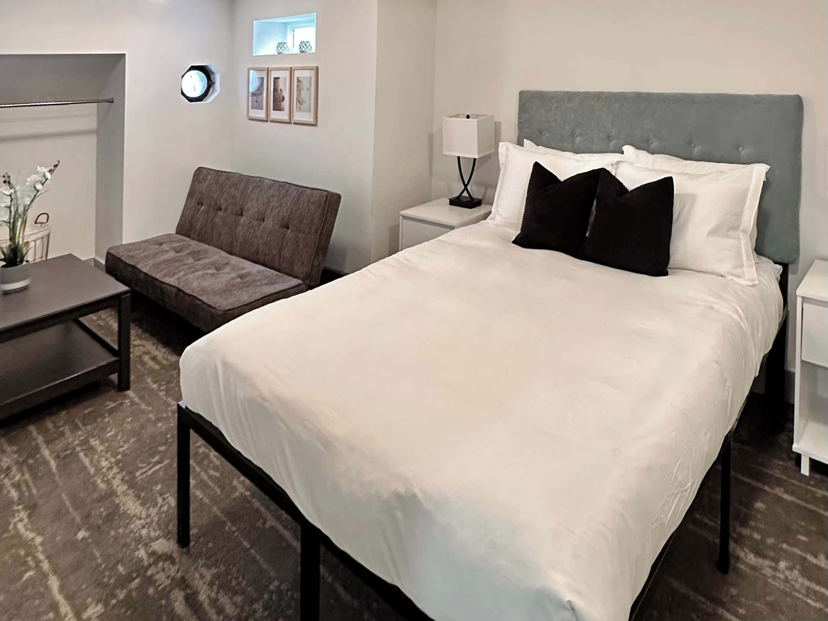 Una habitación de hotel con una cama grande, un sillón tapizado en gris, una mesita y una mesita de noche con lámpara.