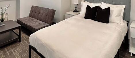 Una habitación de hotel con una cama grande, un sillón tapizado en gris, una mesita y una mesita de noche con lámpara.