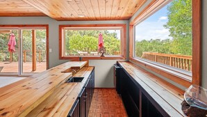 A wooden deck with a kitchen area featuring a sink, cabinets, and a bench. Large windows provide views of greenery.