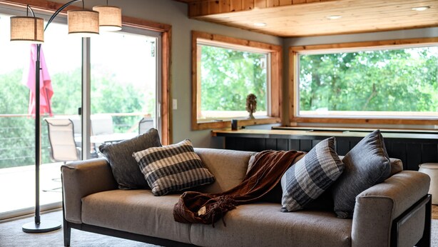 A cozy room with a sofa, a floor lamp, and large windows offering a view of greenery outside.