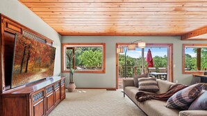 A spacious living room with a wooden ceiling, large windows offering a view of the outdoors, a comfortable sofa with cushions, and a wooden entertainment unit with a flat-screen TV.