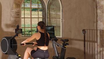 Une personne utilise un appareil d’aviron dans une salle de sport dotée de grandes fenêtres en arc de cercle, offrant une vue sur la verdure à l’extérieur.