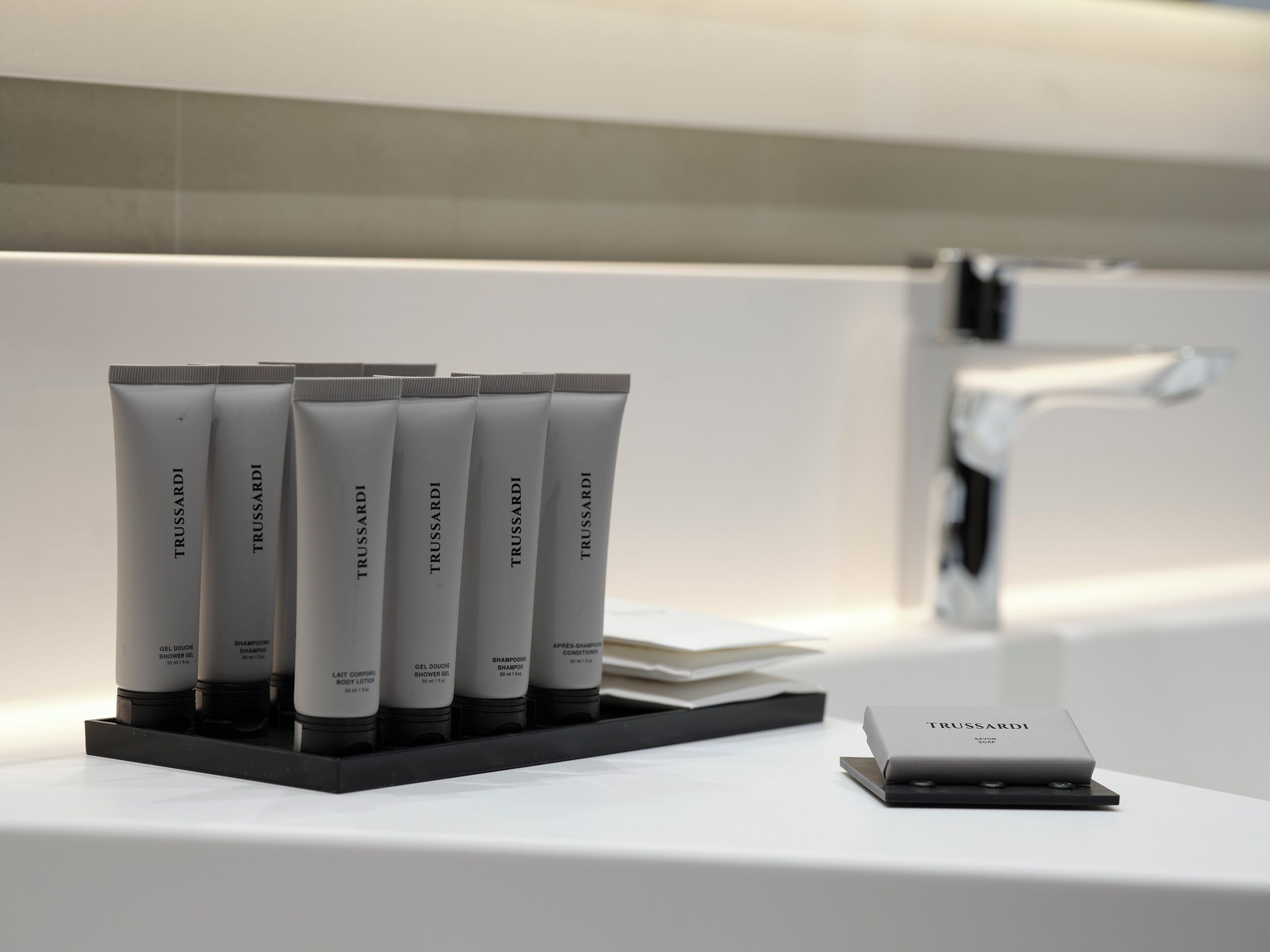 Ein Badezimmer mit einem weißen Waschbecken, einer schwarzen Ablage mit sechs Trussardi-Toilettenartikeln und einer kleinen weißen Schachtel auf der Ablagefläche.