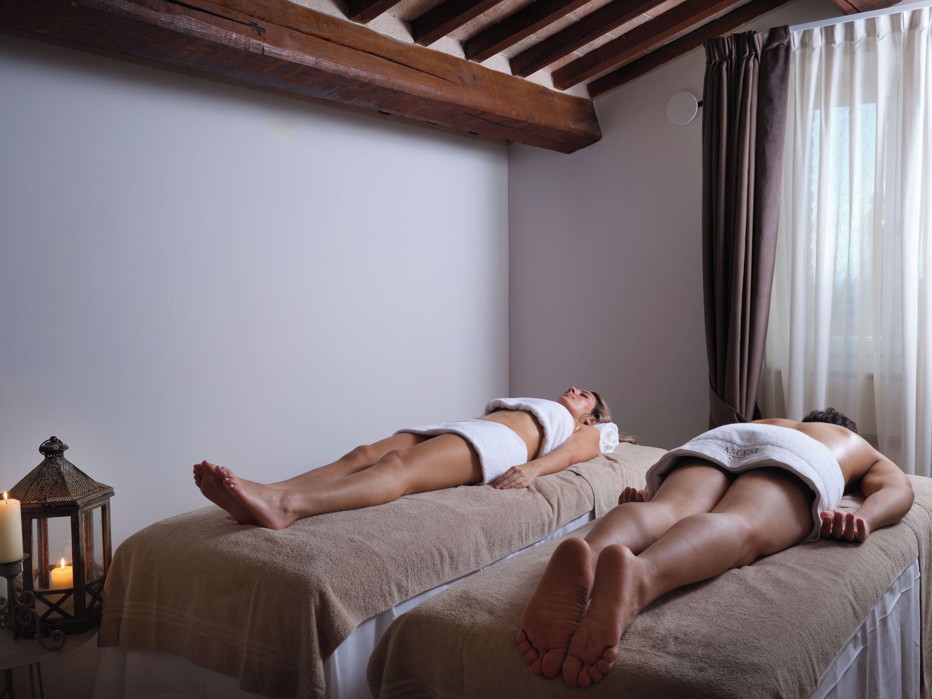 Zwei Massagetische in einem Raum mit Holzboden und einer Kerze auf einem Beistelltisch.