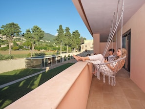 Een balkon met een hangmat, een stenen muur en uitzicht op een groen landschap met bomen en een gebouw.