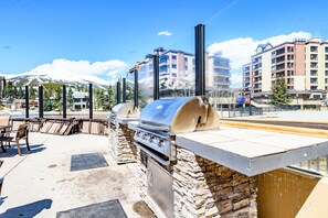 An outdoor barbecue area with stone walls and a grill, overlooking a snowy mountain and buildings in the background.