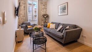 Un salon avec un canapé gris, une petite table noire ornée d’un vase de fleurs, un plancher en bois et un mur en pierre.
