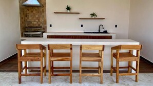 A modern kitchen with a white island, wooden chairs, a stone backsplash, and a hanging light fixture.