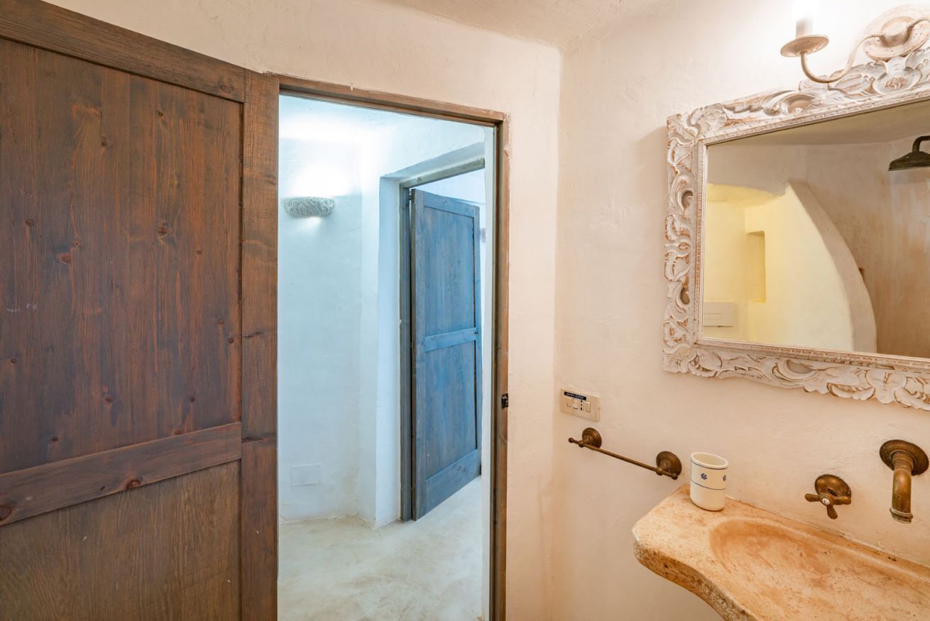 A bathroom with a wooden door, a mirror with an ornate frame, a sink with a vintage faucet, and a light switch on the wall.