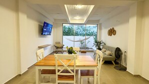 A modern dining area with a wooden table and white chairs, a television mounted on the wall, and a large window offering a view of greenery outside.