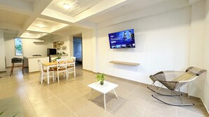 A modern living room with a dining area, a kitchen in the background, and a TV mounted on the wall.