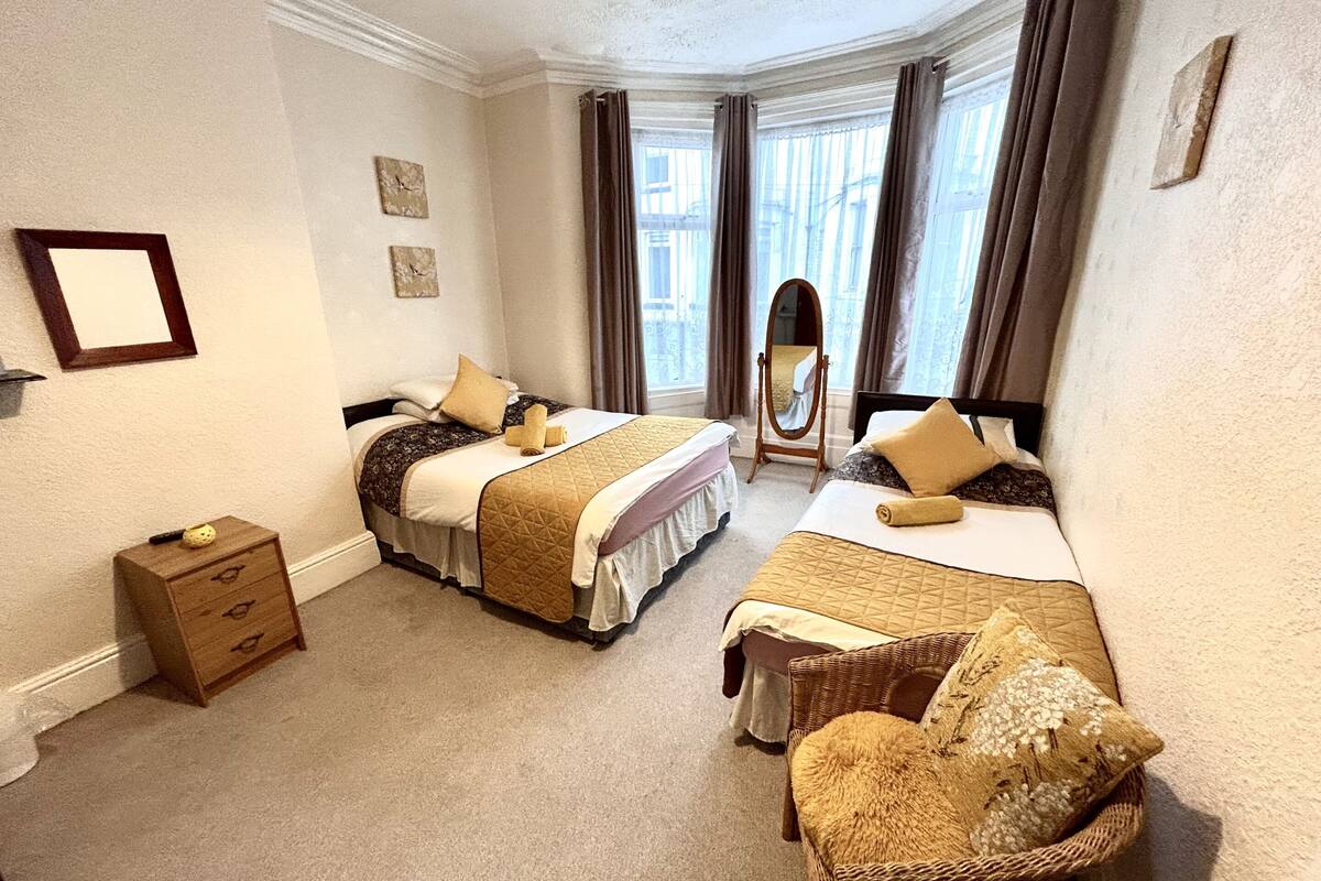 Habitación de hotel con dos camas, un pequeño aparador de madera, una silla de mimbre y un ventanal con cortinas.