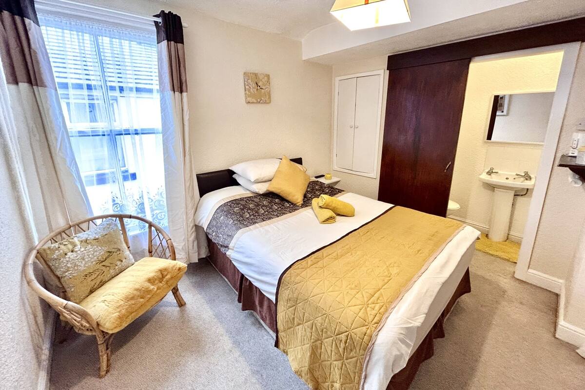 Un dormitorio con cama, una silla, mesita de noche, ventana con cortinas, puerta y baño con lavamanos.
