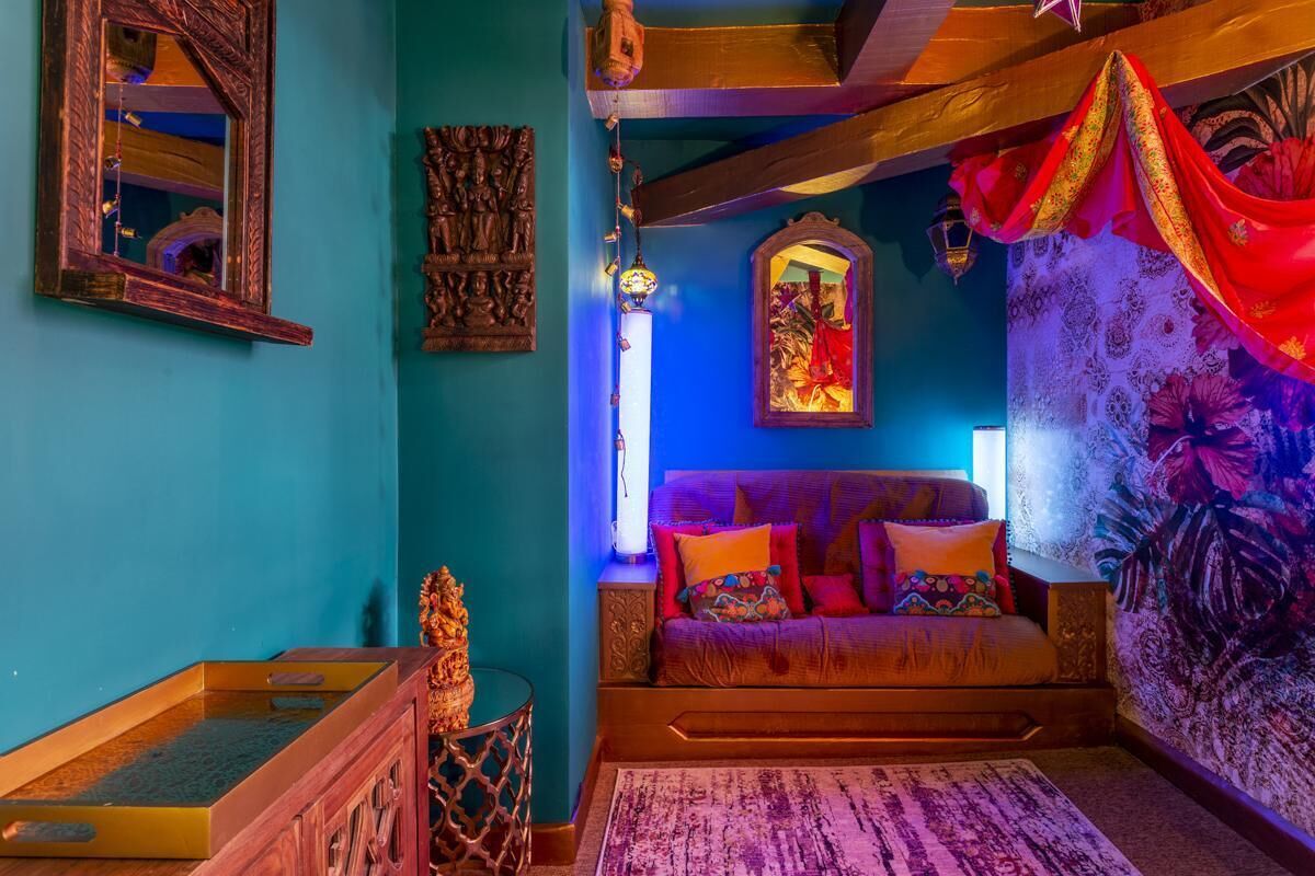 A room with a blue wall, a wooden sofa with colorful cushions, a small table with a decorative item, and a patterned rug.