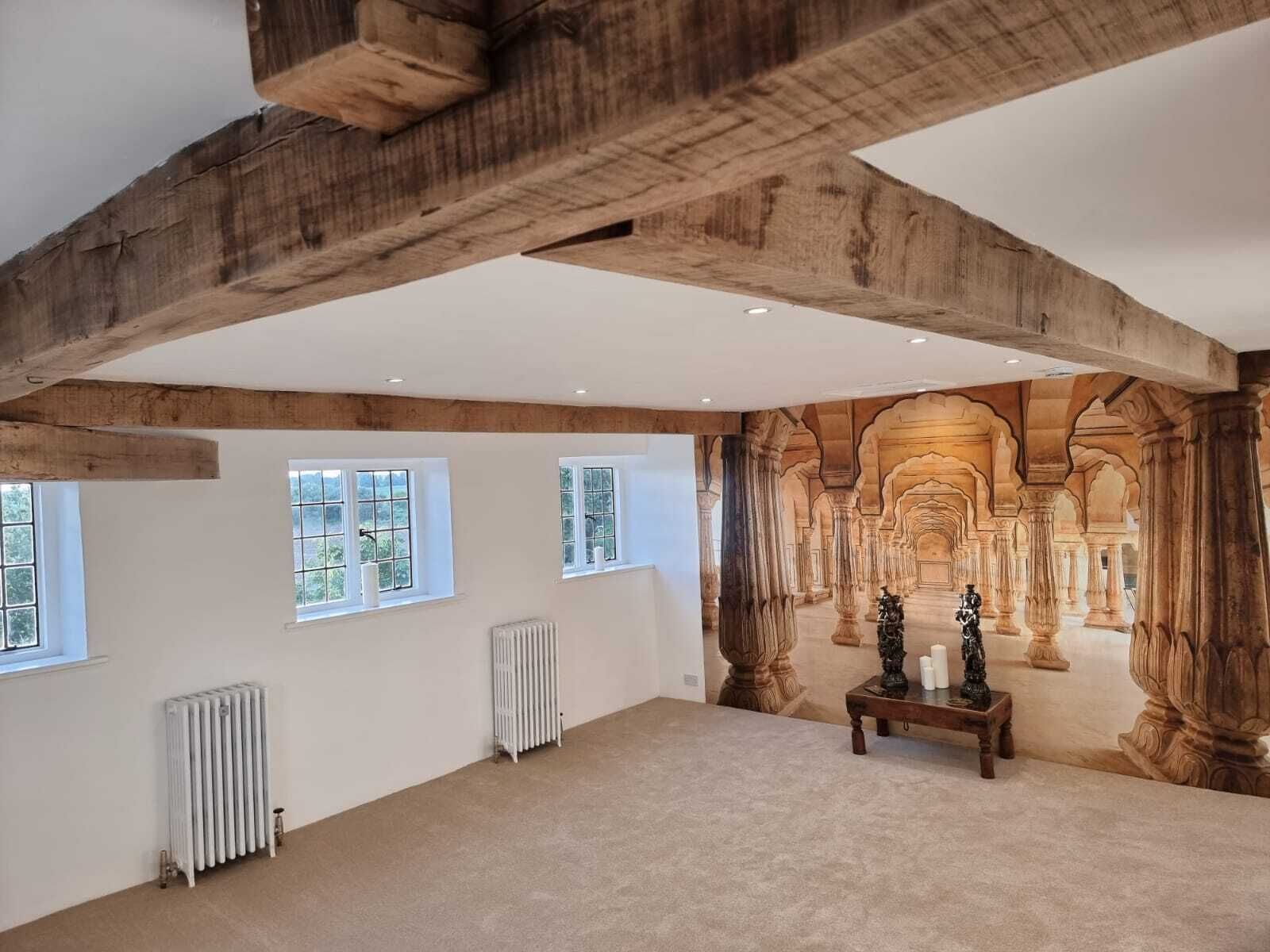Amplia habitación con vigas de madera a la vista, ventanas grandes y un mural que representa un interior arquitectónico ornamentado.