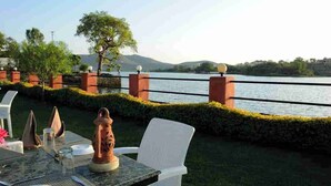 An outdoor dining area with a table set for two, overlooking a body of water and hills. - Pichola Vilas (Udaipur)