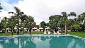 A swimming pool with lounge chairs, palm trees, and thatched-roof huts. - Pichola Vilas (Udaipur)