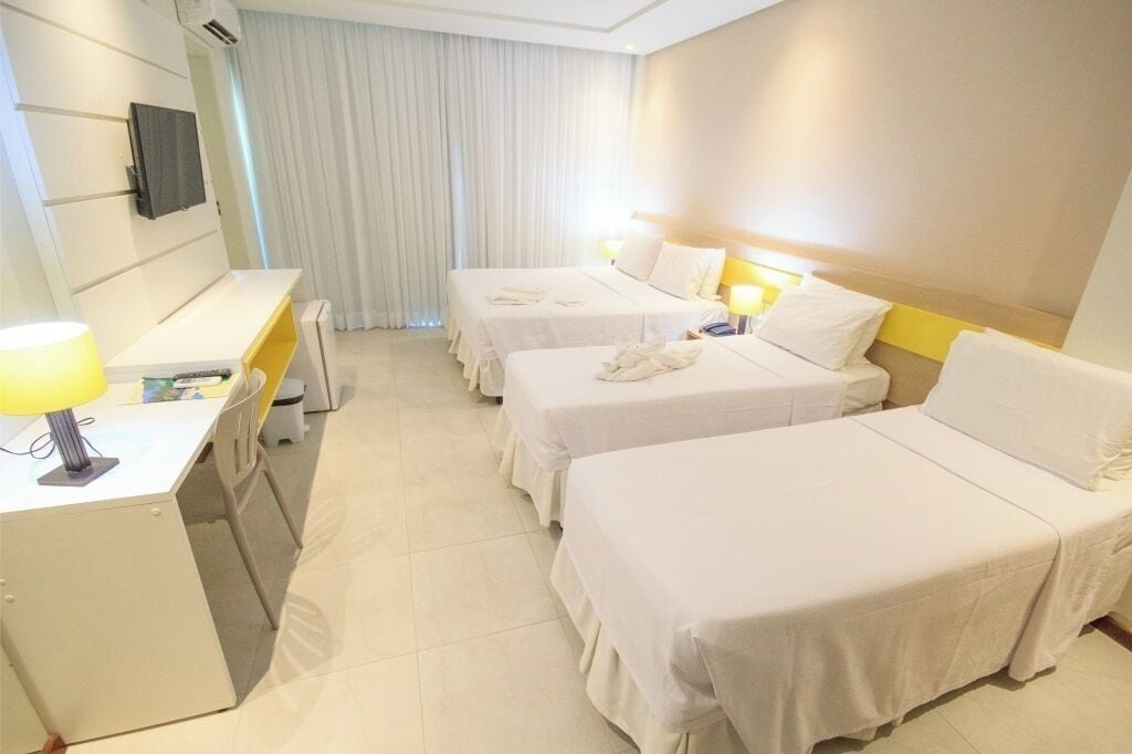 A hotel room with three single beds, a desk with a lamp, and a TV mounted on the wall.