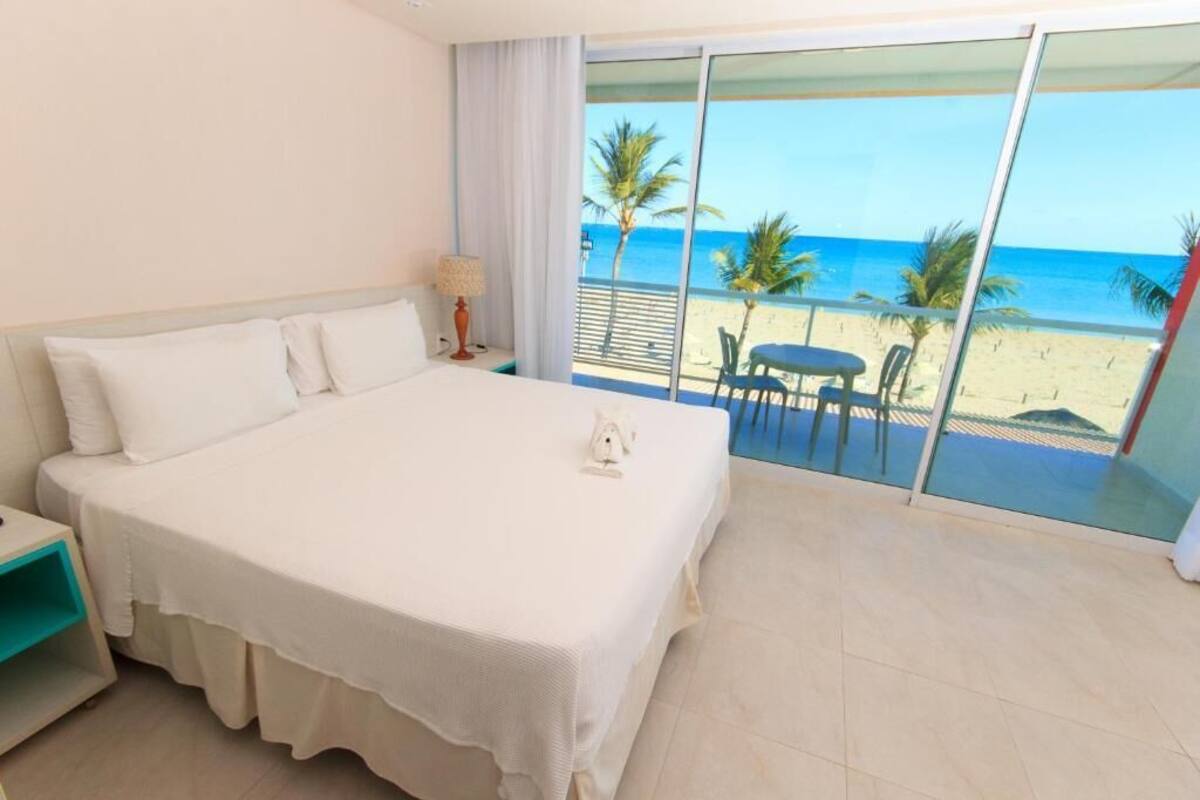 Ein Hotelzimmer mit einem großen Bett, einem Nachttisch, einer Lampe und Blick auf den Strand durch ein großes Fenster.