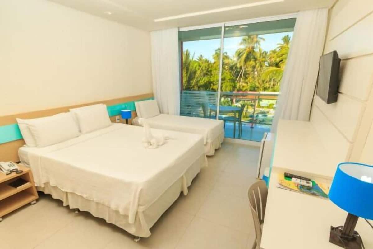 Ein Hotelzimmer mit einem großen Bett, einem kleineren Bett, einem Schreibtisch mit Stuhl, einem Fernseher und einem Balkon mit Blick auf Palmen.
