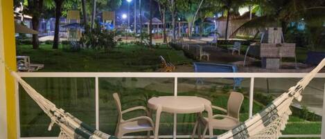 A hammock chair set up on a balcony with a view of a well-lit outdoor area with trees and a pool.