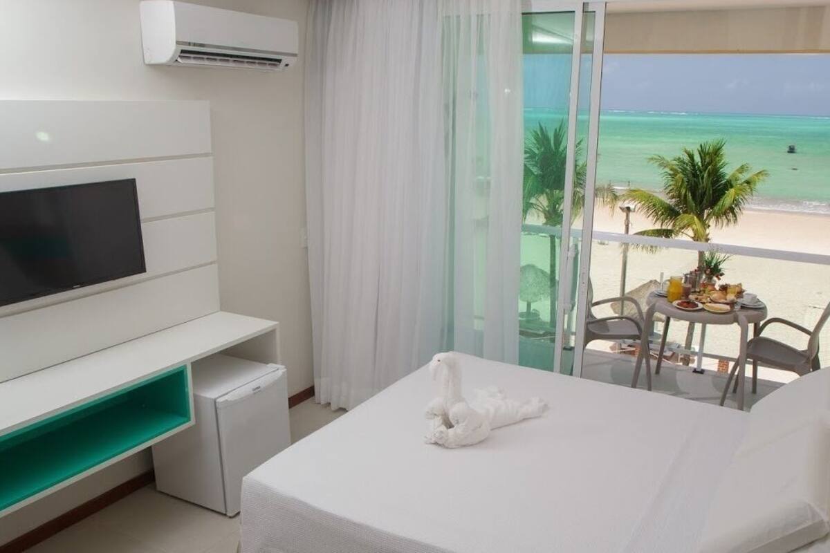 Ein Hotelzimmer mit Bett, Fernseher, einer kleinen Kühlbox, einem Balkon mit Tisch und Stühlen und Blick auf den Strand.