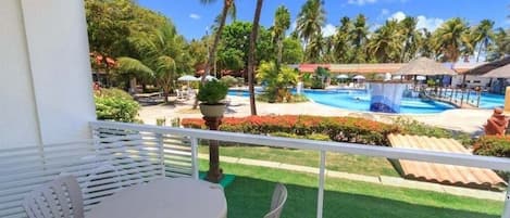 Un balcón con mesa y sillas que da a una piscina rodeada de palmeras y un cielo azul despejado.