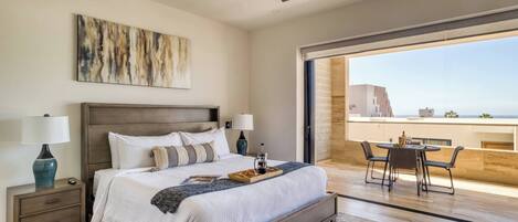 Un dormitorio con cama, mesitas de noche, ventilador de techo, ventana grande y balcón con mesa y sillas.