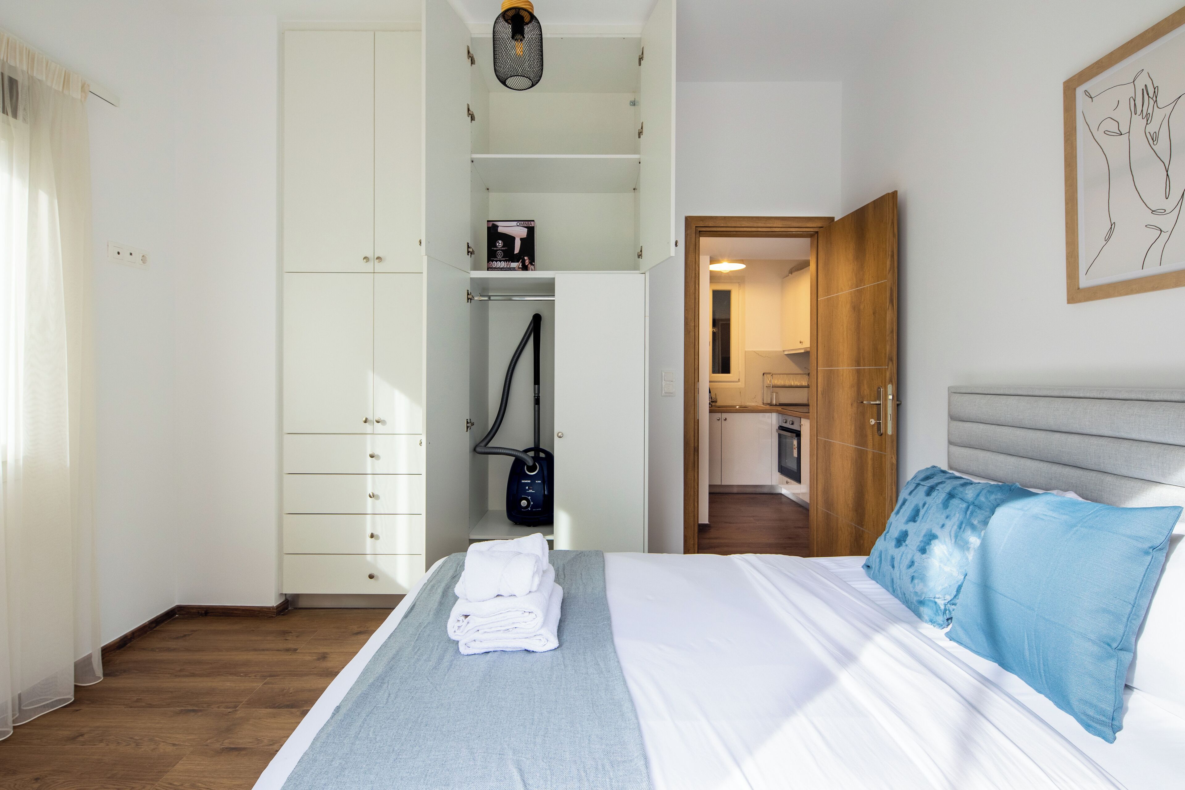 Een slaapkamer met een bed, een kledingkast, een stofzuiger en een keuken op de achtergrond.