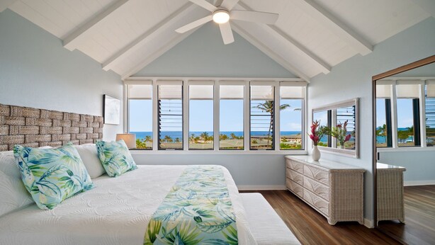 Un dormitorio con una cama grande, un ventilador de techo, vista a la playa y una mesita consola de mimbre.