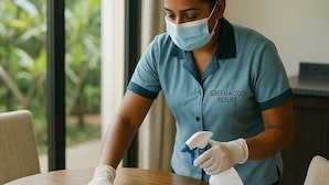 A person cleaning a wooden table with a cloth and spray bottle, wearing a shirt labeled "GREENWOOD RESORT". - Greenwood Udawalawa safari resort (Udawalawa)