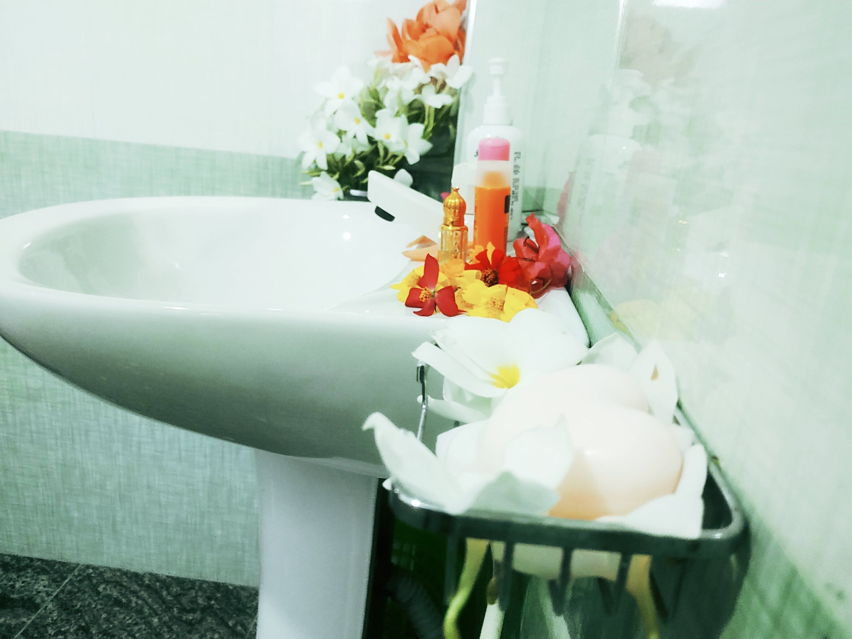 A bathroom sink with a white basin, a small trash bin, and a vase with flowers and a bottle of liquid soap.