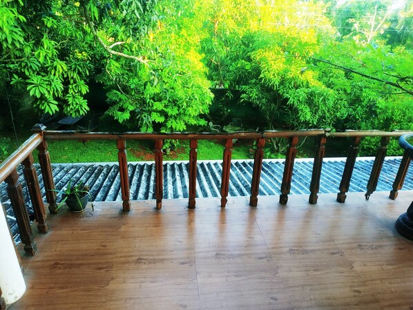 A wooden balcony with a railing overlooking a garden with various plants and a tiled floor.