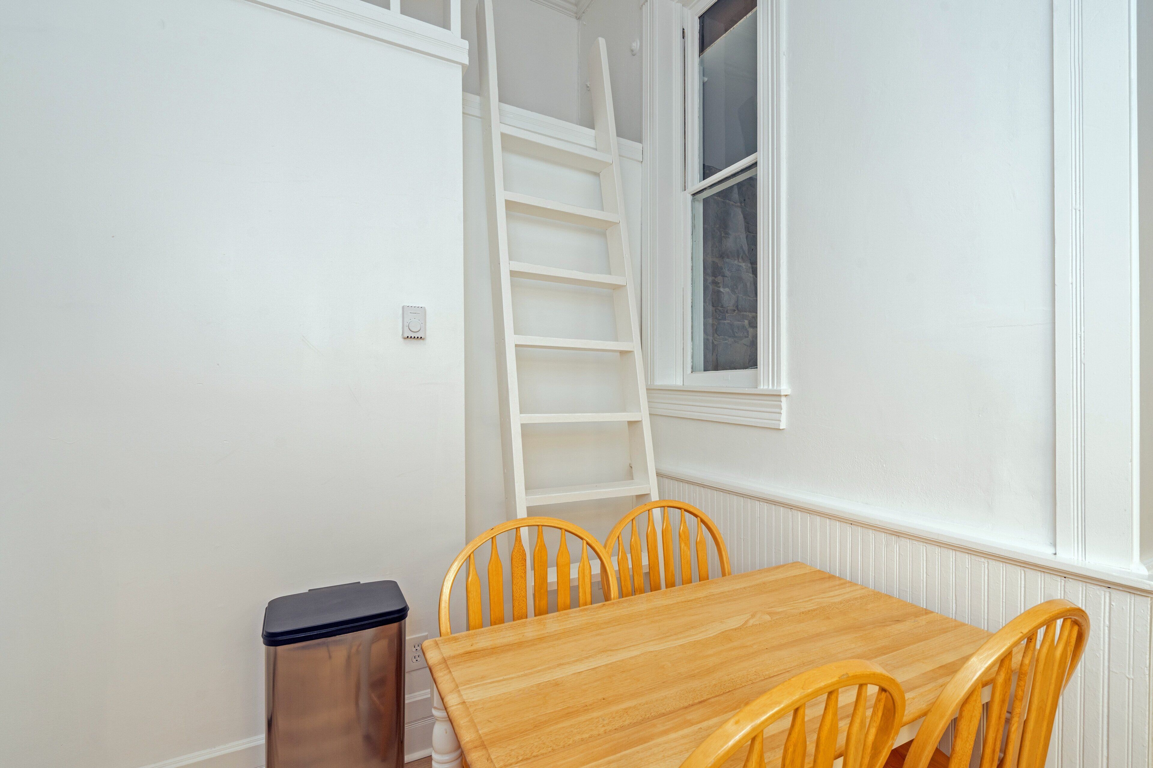 Ein Eckzimmer mit Holztisch und vier Stühlen, einem weißen Heizkörper, einem Fenster mit Steinrahmen und einer weißen, an der Wand lehnenden Leiter.
