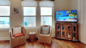 A living room with two wicker chairs, a small round table, a wooden cabinet, and a TV displaying a beach scene. - Casa Blanca | Waterfront, Pool + Pier (Rockport)