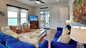 A living room with blue sofas, a coffee table, wicker chairs, a television, and a door leading outside. - Casa Blanca | Waterfront, Pool + Pier (Rockport)