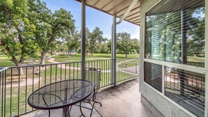 A covered outdoor area with a metal table and chairs, a view of a park with trees and pathways, and a building with large windows. - Ww D101 2 Bedroom Condo (New Braunfels)