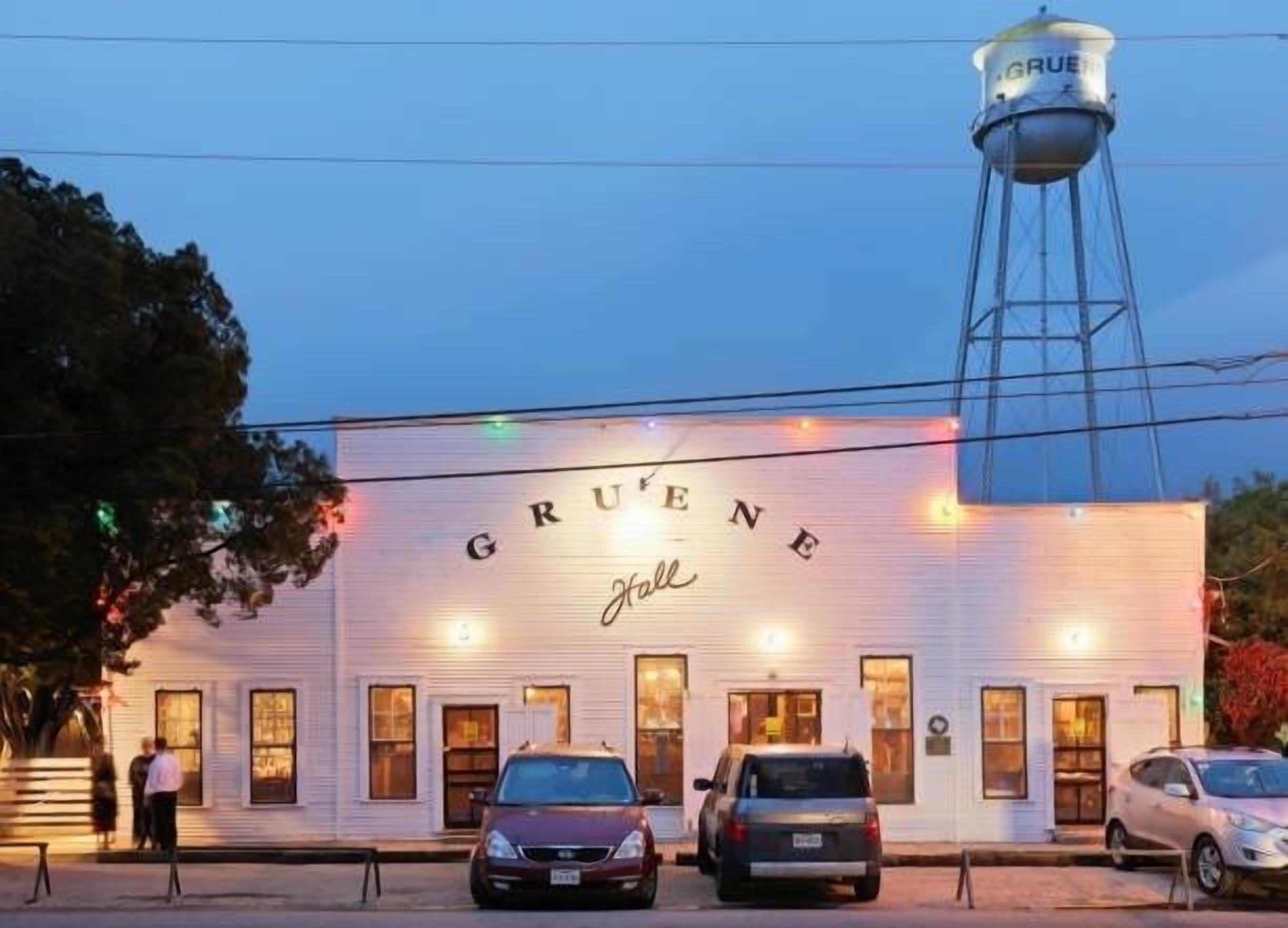 A white building with the name 'Gruene' on the facade, a water tower to the right, and cars parked in front.