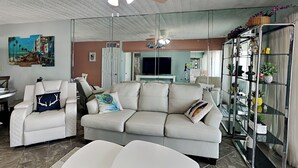 A modern living room with a white sofa, a white armchair, and a glass cabinet with plants. - Sunny Sands | Cozy, Beachfront Condo (Galveston)
