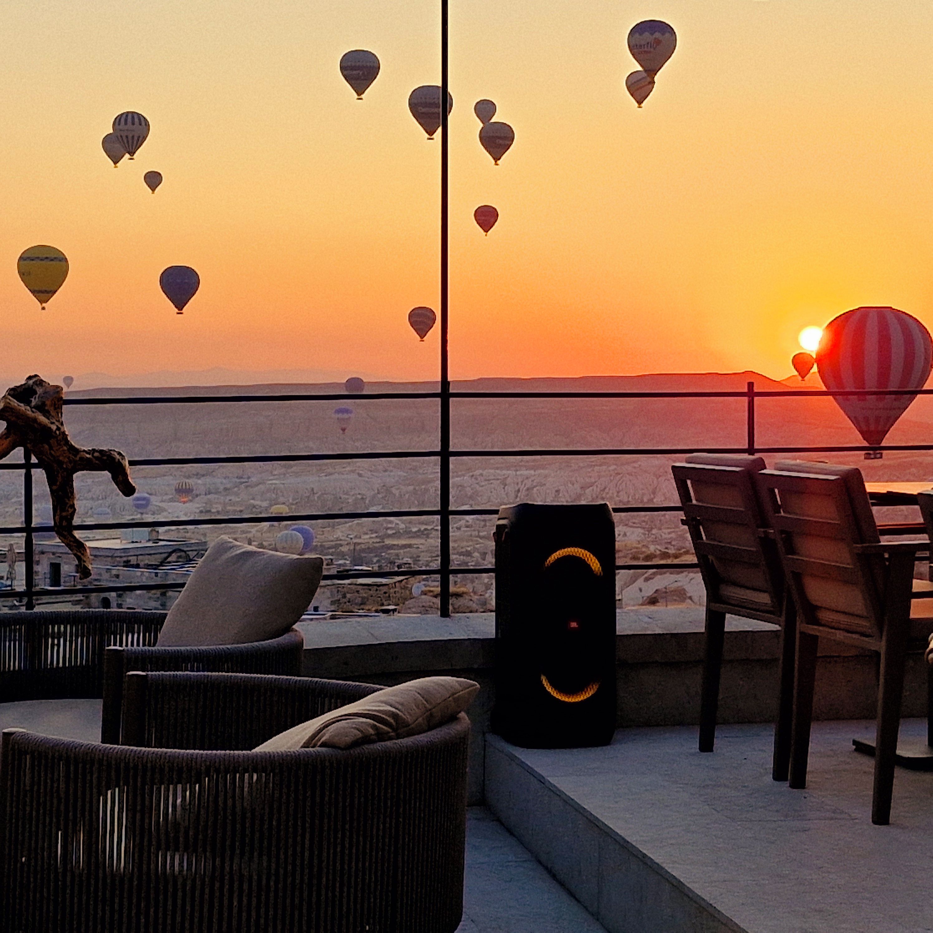 Una terraza con mobiliario de mimbre, un altavoz y vista al mar con globos aerostáticos al atardecer.