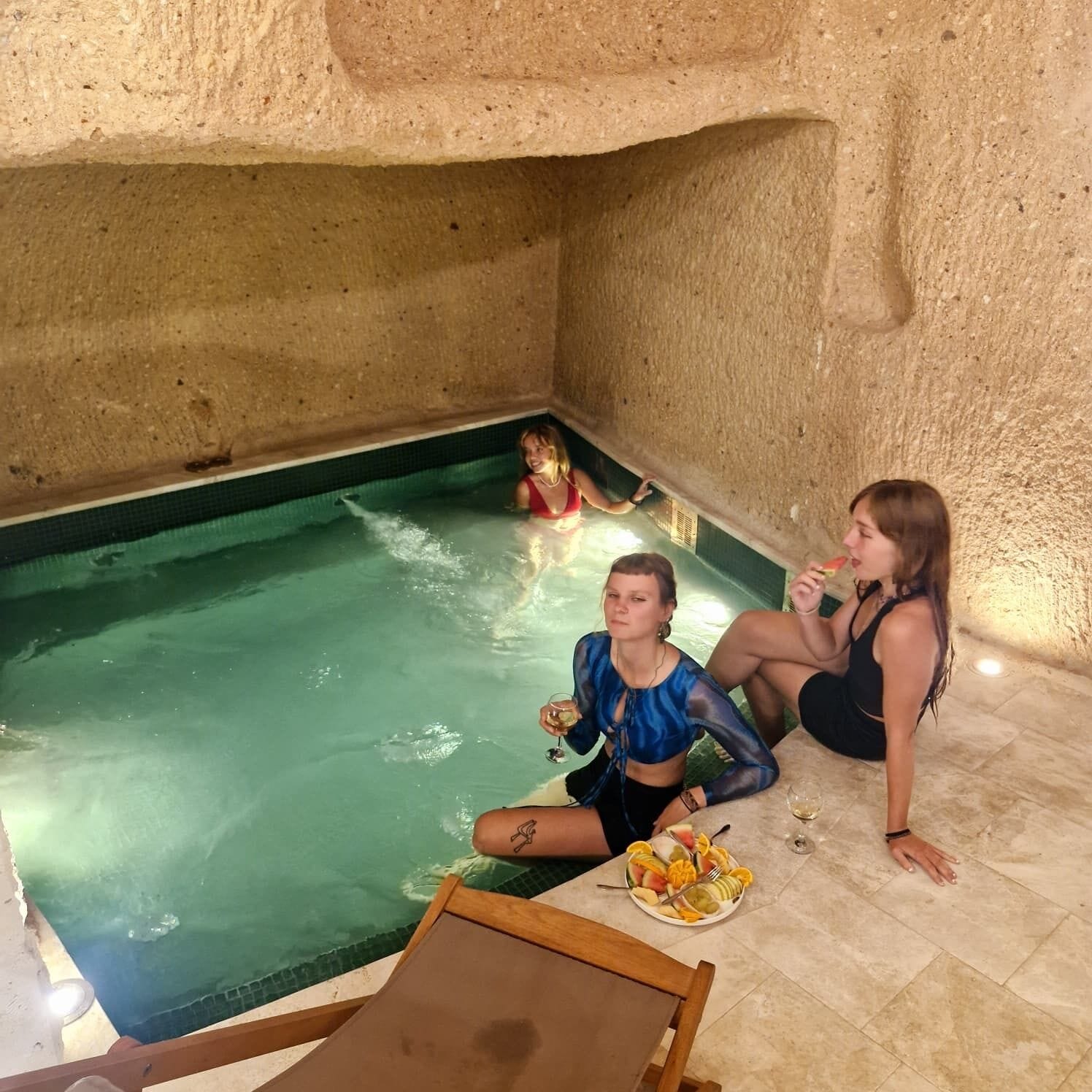 An indoor pool with a stone wall, two people sitting on the floor, and a plate of fruit.