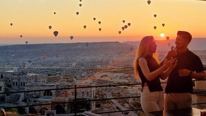 A couple standing on a balcony overlooking a cityscape with hot air balloons in the sky.