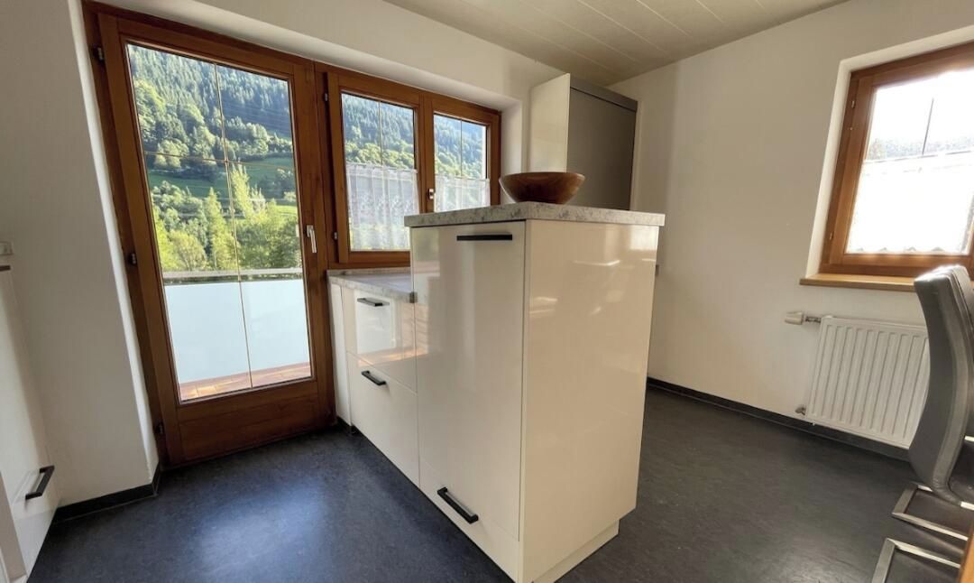A compact kitchen with a refrigerator, a window with a view of greenery, and a door leading to an outdoor area.