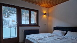 Un dormitorio con techo de madera, una cama con ropa de cama blanca, una ventana con vista a un paisaje nevado y una lámpara fijada a la pared.