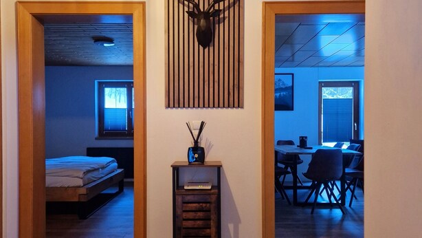 Un interior moderno con techo de madera, una cabeza de venado montada, una mesita con lámpara y un comedor con sillas.