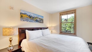 A neatly made bed with white linens, a wooden headboard, and two bedside lamps. A painting of a mountain landscape hangs above the bed, and a window with a view of trees and a building is visible.