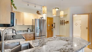 A modern kitchen with a stainless steel refrigerator, marble countertops, wooden cabinets, and pendant lighting.