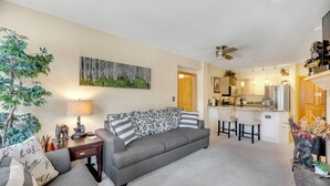A modern living room with a grey sofa, a coffee table, and a lamp. There is a kitchen area with a refrigerator and a bar counter.