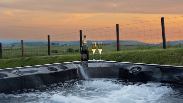 A hot tub with two glasses of white wine and a bottle, surrounded by a grassy field and a wire fence, with a sunset in the background. - Golygfa Cwrt (Talybont)